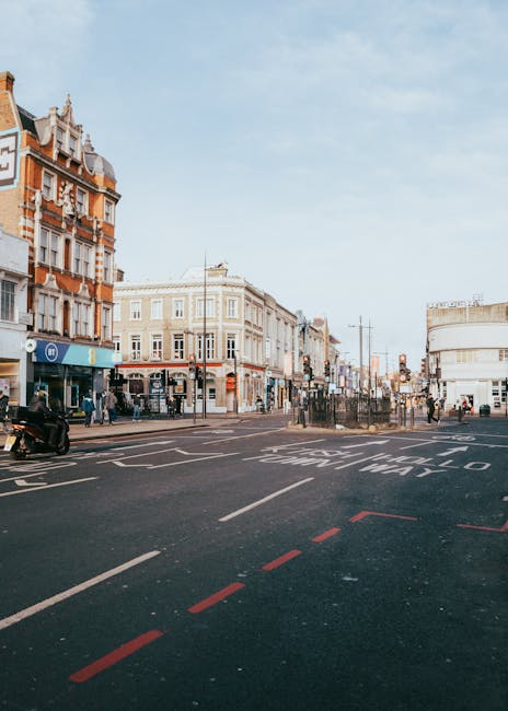 A street scene in Camden Town showing a row of Victorian-style brick buildings with decorative facades, retail shops, and cafés on the ground level. The pavement features pedestrian crossings and road markings, with a few people walking along the sidewalk and some waiting at the crossing. The sky is partly cloudy with soft lighting illuminating the scene. The image captures the lively atmosphere of Camden Town, highlighting its architectural details and urban layout, consistent with the focus on cleaning and maintenance for residential and commercial spaces. For professional cleaning services like surface cleaning, deep cleaning, and sanitisation, Camden Town Cleaners offers expert assistance in keeping such areas tidy and hygienic.