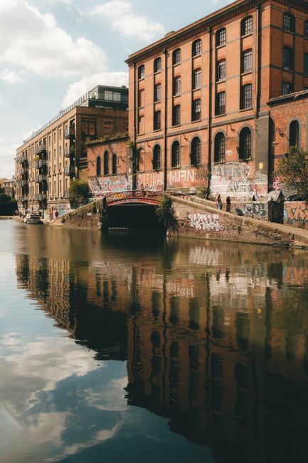A view of a river with calm, reflective water running beneath a small stone bridge in Camden, London. On one side, there is a large red-brick industrial building with multiple arched windows and graffiti-covered walls, indicative of a historic warehouse or factory. Adjacent modern buildings with glass facades line the street, with trees and urban street furniture visible along the pathway. The sky is partly cloudy with sunlight illuminating the scene, emphasizing the textures of the brickwork and the gentle ripples on the water's surface. This urban riverside scene highlights a blend of historic and contemporary architecture in Camden, with reflections creating a symmetrical visual effect on the water. Camden Town Cleaners specializes in surface cleaning, deep cleaning, and sanitisation services for such commercial properties, ensuring hygiene and restored appearance in line with their offerings for the Camden Chalk Farm NW1 area.