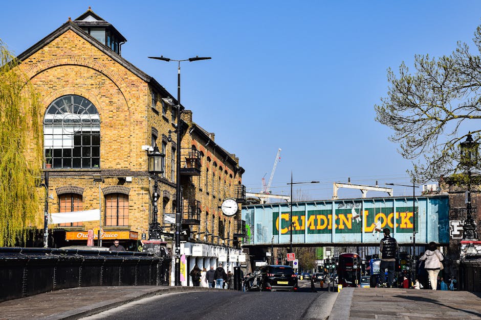 A street scene in Camden Town showing a row of Victorian-style brick buildings with decorative facades, retail shops, and cafés on the ground level. The pavement features pedestrian crossings and road markings, with a few people walking along the sidewalk and some waiting at the crossing. The sky is partly cloudy with soft lighting illuminating the scene. The image captures the lively atmosphere of Camden Town, highlighting its architectural details and urban layout, consistent with the focus on cleaning and maintenance for residential and commercial spaces. For professional cleaning services like surface cleaning, deep cleaning, and sanitisation, Camden Town Cleaners offers expert assistance in keeping such areas tidy and hygienic.