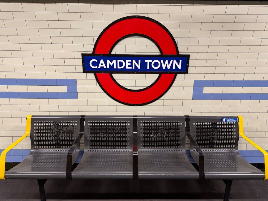 A row of four metal bench seats with perforated surfaces and black frames, located in an underground station with white tiled walls featuring blue horizontal stripes. The back wall displays a large re