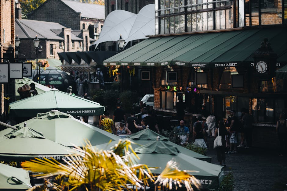Outdoor view of Camden Market with multiple green umbrellas covering outdoor seating areas, wooden dining tables, and a diverse crowd of people walking and socializing. The market buildings feature traditional brick facades with green awnings labeled 'Camden Market' and a clock mounted on one of the structures. In the background, modern glass and steel structures are visible, along with a white domed roof. The scene is illuminated by natural daylight, highlighting the clean and well-maintained surfaces of the umbrellas, tables, and surrounding architecture. The area appears lively and bustling, reflecting a vibrant urban environment typical of Camden Town, where surface cleaning and maintenance by Camden Town Cleaners contribute to its welcoming atmosphere.