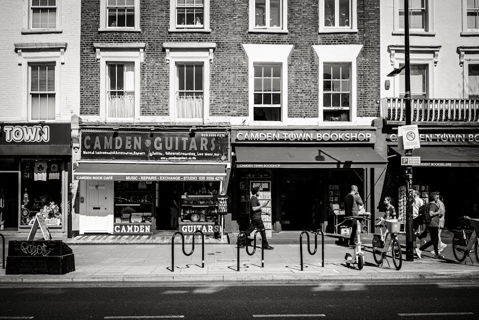 A black-and-white photograph of a street scene in Camden Town, featuring storefronts including Camden Guitars and Camden Town Bookshop with large signage. The buildings have brick facades with multiple rectangular windows. The sidewalk is occupied by pedestrians, some carrying bags or pushing bicycles, and a few are engaged in conversation. The street has metal bike racks and a lamppost, with a no-parking sign attached. The scene is evenly lit, capturing the urban atmosphere with clean, well-maintained surfaces typical of a commercial area in Camden Town. Camden Town Cleaners is a local cleaning company that offers surface cleaning, deep cleaning, and sanitisation services, ensuring hygienic environments in residential and commercial spaces.