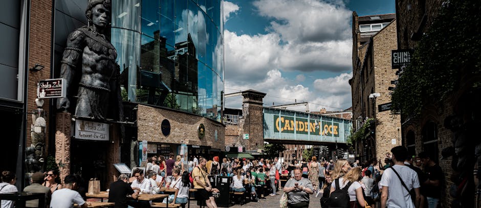 A busy outdoor area in Camden Town with numerous people seated at wooden tables on a paved surface, surrounded by brick buildings and modern glass-fronted shops. Visible surfaces include concrete or stone pavement, with no apparent dirt or debris. The scene features a prominent sign reading 'Camden Lock' hanging over the street. The lighting is natural daylight, casting soft shadows, and the scene appears clean and well-maintained. Camden Town Cleaners' focus on surface cleaning and deep sanitisation aligns with ensuring such vibrant public spaces remain hygienic and inviting for visitors.