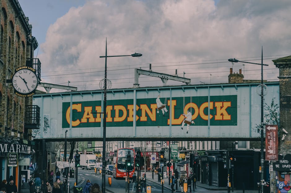 An outdoor view of Camden Lock in Camden Town, featuring a large rectangular sign with the words 'CAMDEN LOCK' painted in bold yellow and orange letters on a green background, mounted on a metal structure spanning the street. In the foreground, a busy urban scene includes pedestrians walking along the pavement, a red double-decker bus, and small shops with signage. The sky above is partly cloudy with soft light illuminating the scene, and vintage-style street lamps and overhead wires are visible against the backdrop of brick buildings. The overall image captures the vibrant atmosphere of this popular area, emphasizing the lively street environment near Camden Lock, a focal point for visitors and locals alike.